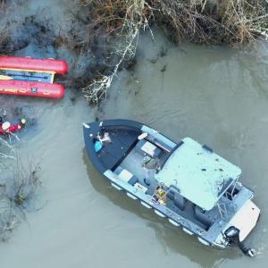 Overhead photo of a red rescue boat floating in a muddy river channel choked with branches and debris.