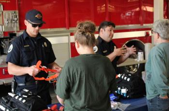 Firefighter showing off equipment to a visitor with a red ambulance in the background.