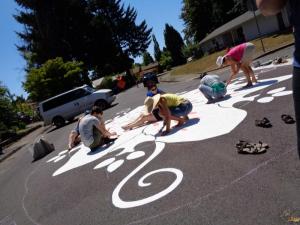 Volunteers painting a mural on the street outside on a sunny day. A large tree provides shade in the background.