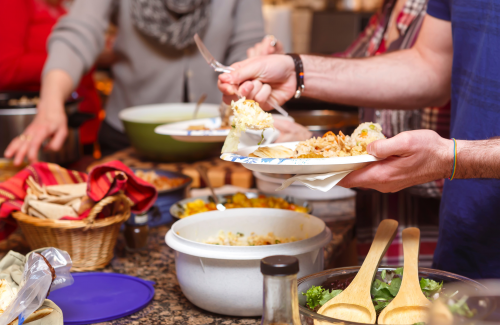 potluck table of food, person wearing rainbow bracelet fills a plate