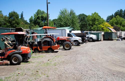 Orange tractors and lawnmowers parked on a gravel lot on a sunny day with blue sky and green trees in the background.