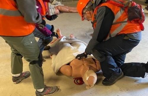 Volunteers wearing orange vests conducting first aid drills on a dummy lying on the ground.