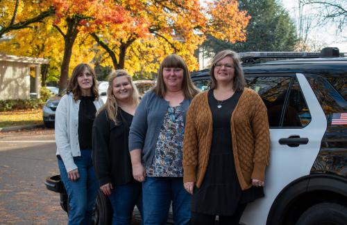 Four women stand in front of a police cruiser outside in the fall.