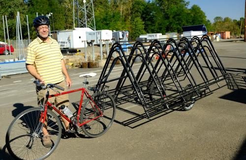 Cyclist standing outside on a gravel parking lot next to a bike with a long trailer, upon which is loaded 3 or 4 bike racks.