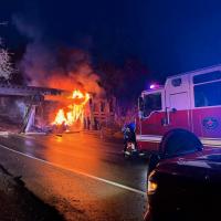 Wooden railroad bridge on fire at night with fire engine in the foreground and firefighters using hoses.