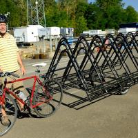 Cyclist standing outside on a gravel parking lot next to a bike with a long trailer, upon which is loaded 3 or 4 bike racks.
