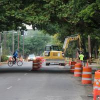 Bicyclist riding across a street under construction, with orange barrels and excavation vehicles working in the roadway.