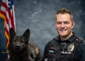 K9 Nexus, a large dark dog, and Officer Diebel in front of a blue background and American flag