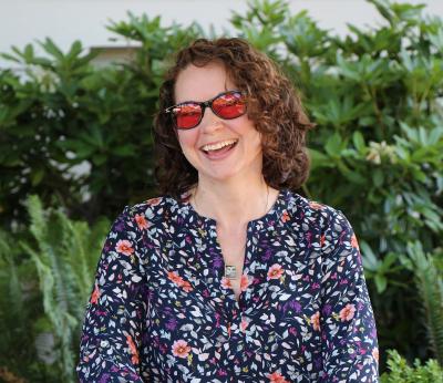 Headshot of Named City Employee with short curly hair, wearing a blue floral print top and rose-tinted glasses, and is smiling.