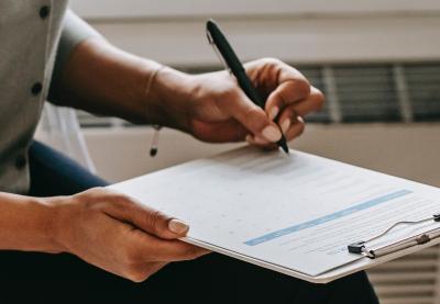 Person seated near a heat register filling out a form using a pen and a clipboard.