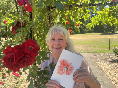 Community Member Cathy Pettigrew holds a painting of a rose next to a rose bush at the Avery Park Rose Garden