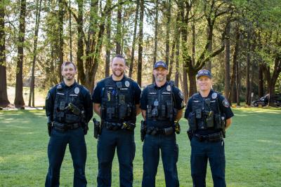 CLU Group of four uniformed police officers stand in a park with large trees in the background