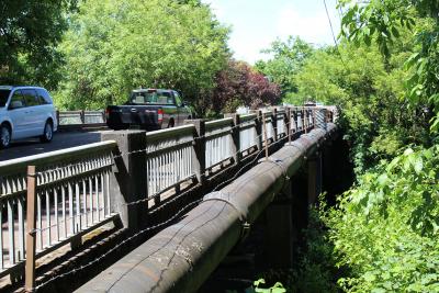 Water pipe running alongside a bridge with green trees and bushes nearby.