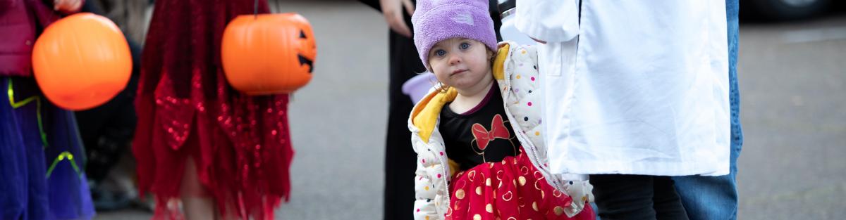 Young child dressed up like minnie mouse peaking out from behind an adult's leg, halloween pumkin baskets in the background