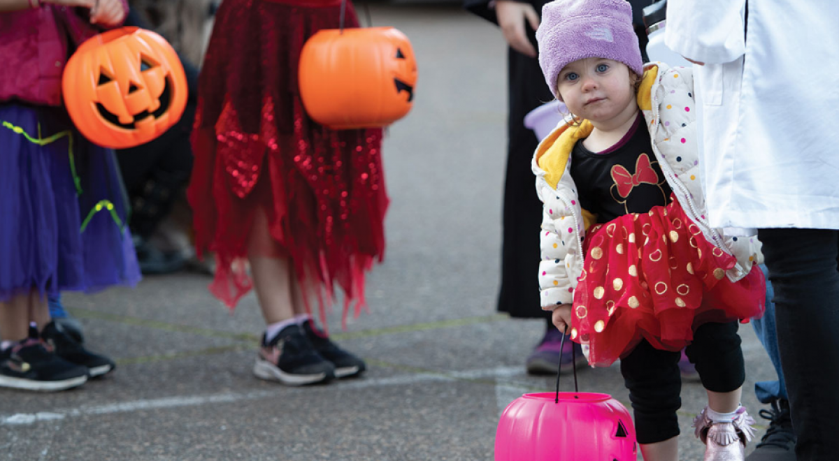 little kid dressed in halloween costume and coat looking at the camera while holding a pink jack-o-lantern