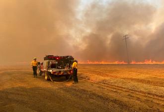 Corvallis firefighters with a small fire engine in a dry grass field, with a line of bright orange flames in the background