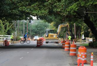 Bicyclist riding across a street under construction, with orange barrels and excavation vehicles working in the roadway.