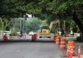 Bicyclist riding across a street under construction, with orange barrels and excavation vehicles working in the roadway.